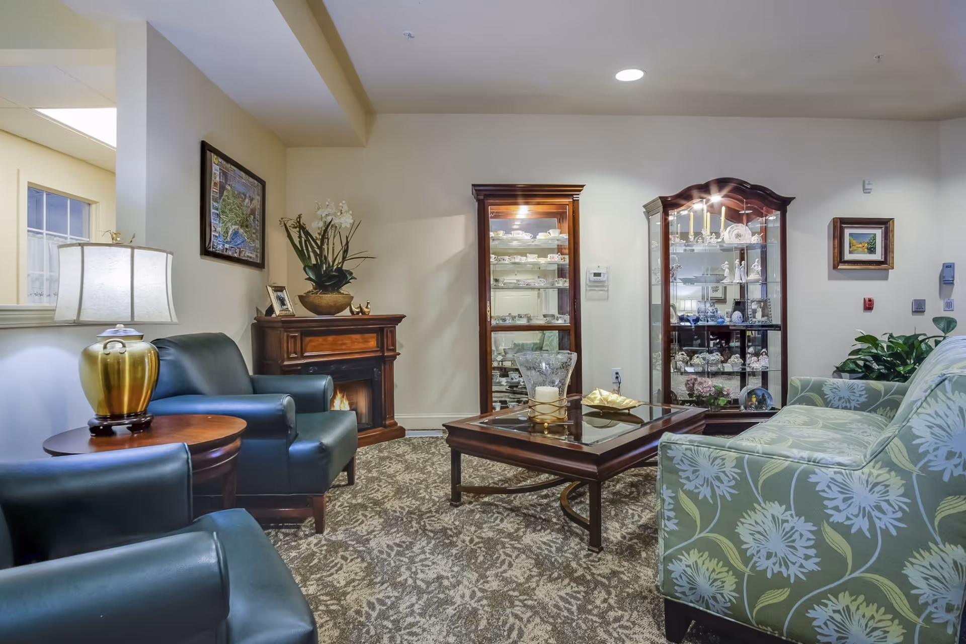 A cozy living room area with two dark green armchairs, a floral patterned sofa, a wooden coffee table with decorative items, a small electric fireplace, two glass display cabinets filled with china and figurines, a table lamp, and framed artwork on the walls.