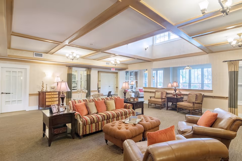 A spacious and well-lit living room area in a senior living facility featuring a striped sofa with multiple cushions, two leather armchairs with orange pillows, a tufted ottoman with books on top, side tables with lamps and framed photos, and additional seating with armchairs and a small table near large windows. The room has a coffered ceiling with chandeliers and beige walls with curtains on the windows.