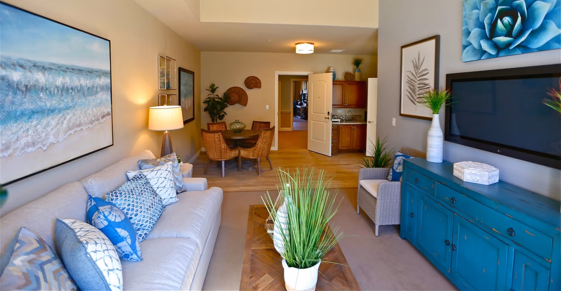 A cozy living room with a beige sofa adorned with blue and white patterned pillows, a wooden coffee table with green plants, a wicker armchair, and a blue cabinet with a flat-screen TV mounted above it. The room features beach-themed artwork on the walls and a small dining area with a round table and four wicker chairs in the background. The flooring is a combination of carpet and wood, and there is a small kitchenette visible through an open door.