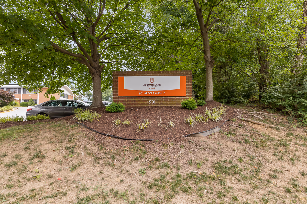 Outdoor view of a brick sign for Autumn Lake Healthcare at Arcola, located at 901 Arcola Avenue, surrounded by trees and landscaping with a parked car and building partially visible in the background.
