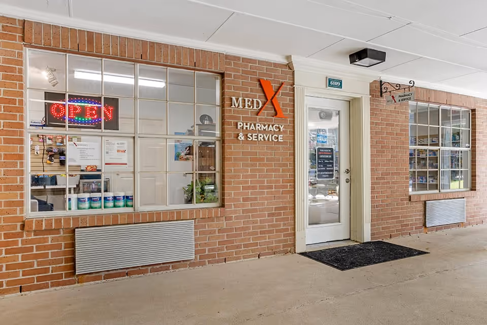 Brick storefront of MedX Pharmacy & Service with large display windows, an 'OPEN' neon sign, and a glass door under a covered walkway.