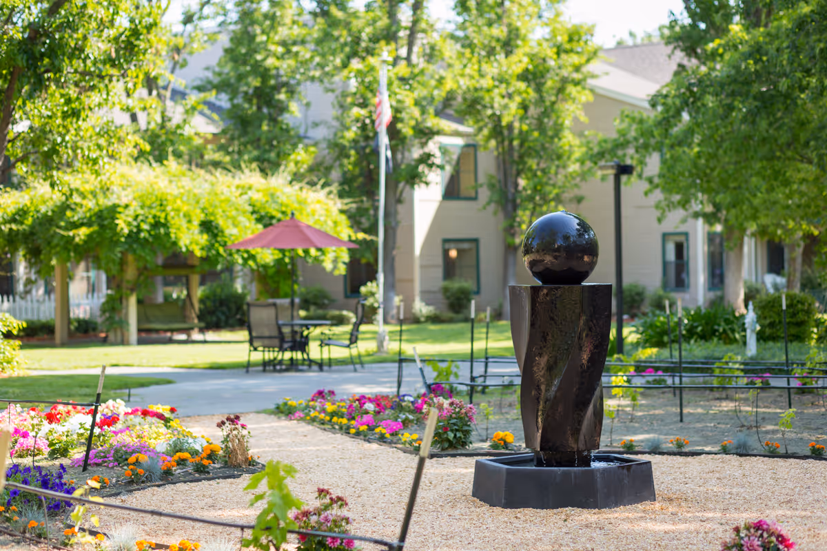 A peaceful outdoor garden area at Cogir of Vacaville featuring a modern black water fountain with a spherical top, surrounded by colorful flower beds and gravel pathways. In the background, there are green trees, a table with chairs under a red umbrella, and a beige building with green-trimmed windows.