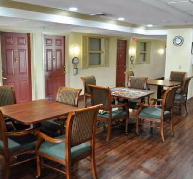 Interior view of a common area in a senior living facility with several wooden tables and chairs arranged on a polished wooden floor. The walls are light-colored with red doors and small windows with shutters. Wall-mounted lights and a clock are visible on the walls.
