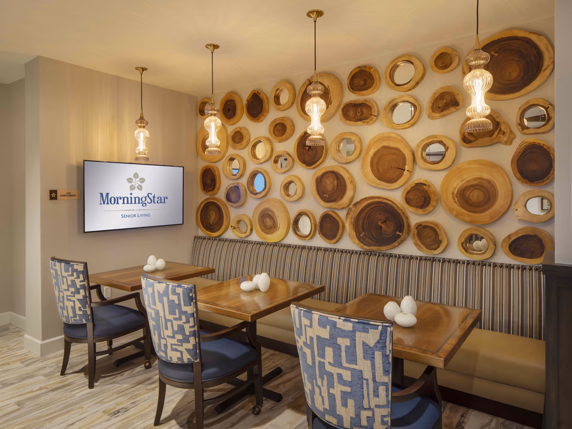A cozy dining area in MorningStar Assisted Living & Memory Care of Rio Rancho featuring two wooden tables with blue patterned chairs and a striped bench along the wall. The wall is decorated with circular wooden slices and mirrors, and three pendant lights hang from the ceiling. A screen on the wall displays the MorningStar Senior Living logo.