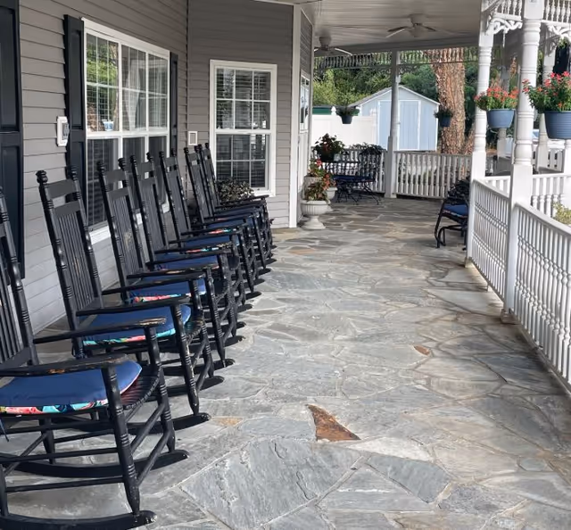 A long covered porch with a row of black rocking chairs with blue cushions lined up against the wall of a gray building. The porch has a stone floor and white railing, with potted plants and hanging flower pots. A white shed and trees are visible in the background.