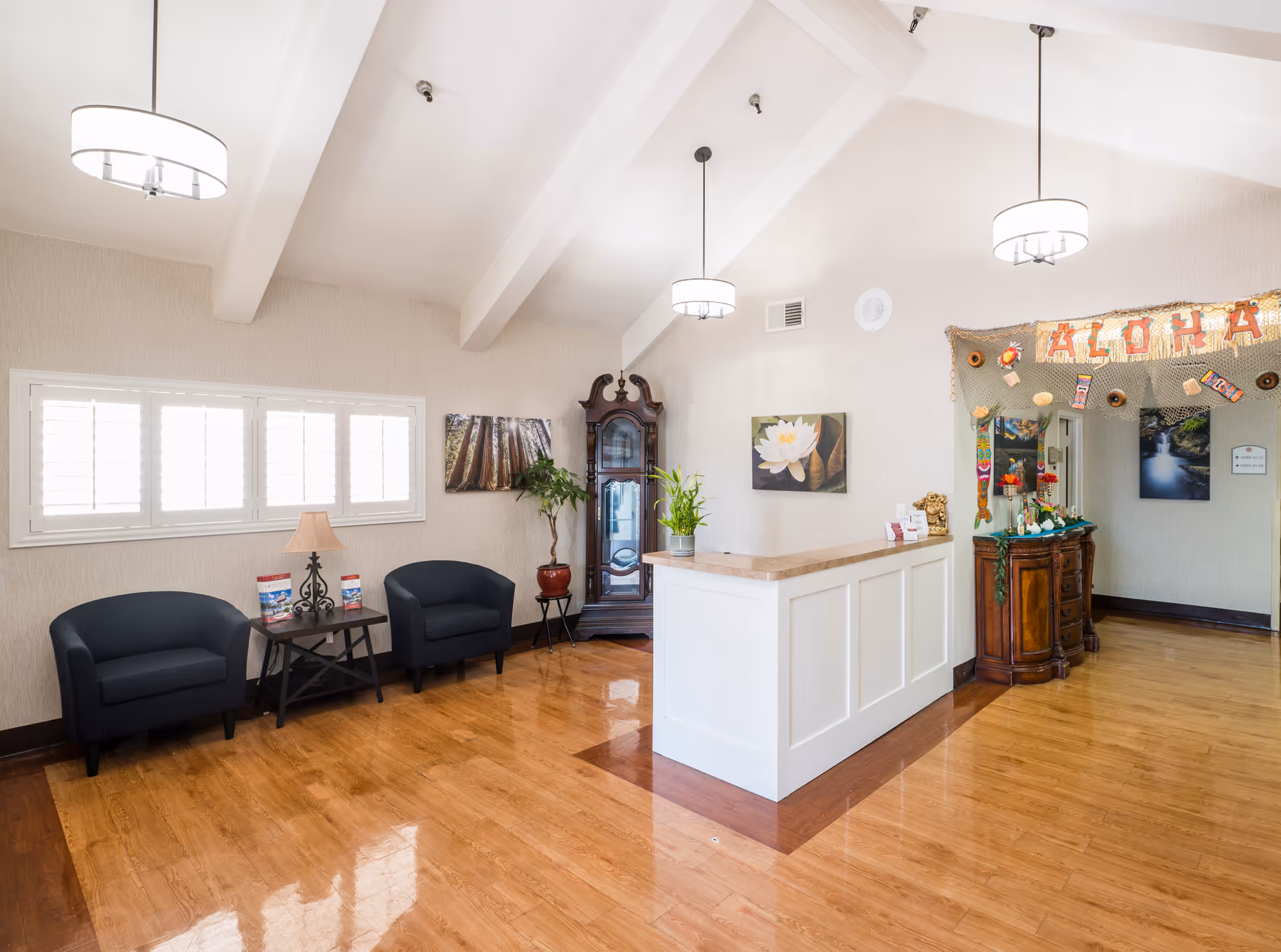 A bright and clean reception area with a white reception desk, two black armchairs with a small table and lamp between them, a tall wooden grandfather clock, and decorative plants. The walls are light-colored with framed artwork, and there is a festive 'ALOHA' banner hanging above a wooden sideboard. The floor is polished wood, and three modern pendant lights hang from the vaulted ceiling.