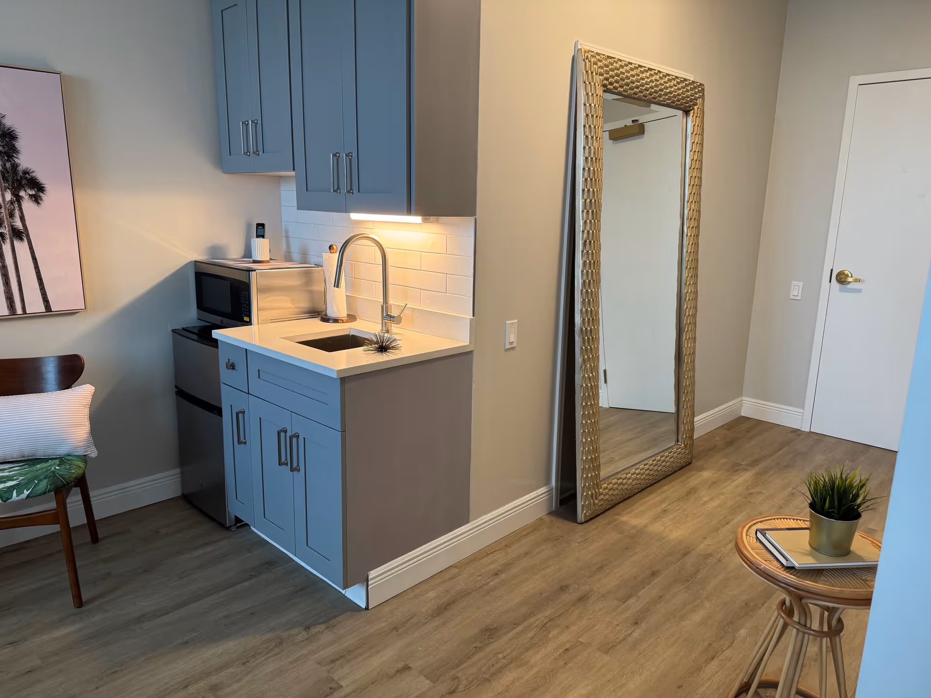 A small kitchenette area with light blue cabinets, a stainless steel microwave and mini fridge, a sink with a modern faucet, and a paper towel holder. To the right, there is a large decorative gold-framed mirror leaning against the wall. The floor is wood, and there is a small round table with a plant and books on it. A chair with a cushion and a wall art featuring palm trees are partially visible on the left side.