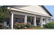 Front exterior view of Lighthouse Rehab & Healthcare building with white columns, a covered porch, rocking chairs, and landscaping including bushes and flowers under a clear blue sky.