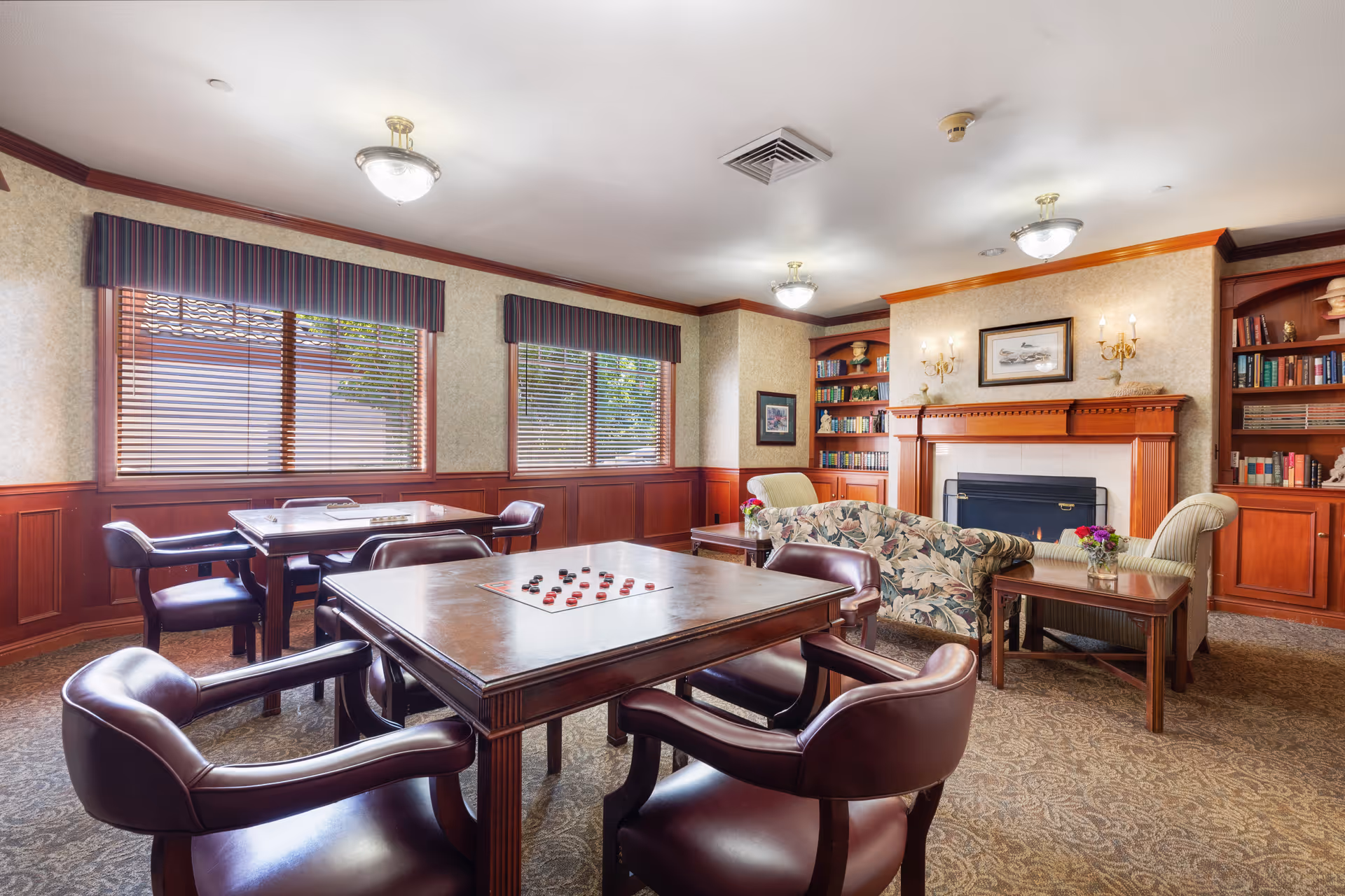 A cozy common room with two tables and four leather chairs each, one table set up for a board game. The room features large windows with blinds and valances, a fireplace with a wooden mantle, built-in bookshelves filled with books and decorative items, and comfortable upholstered chairs and a floral-patterned sofa. The walls have wood paneling and the carpet has a subtle pattern.