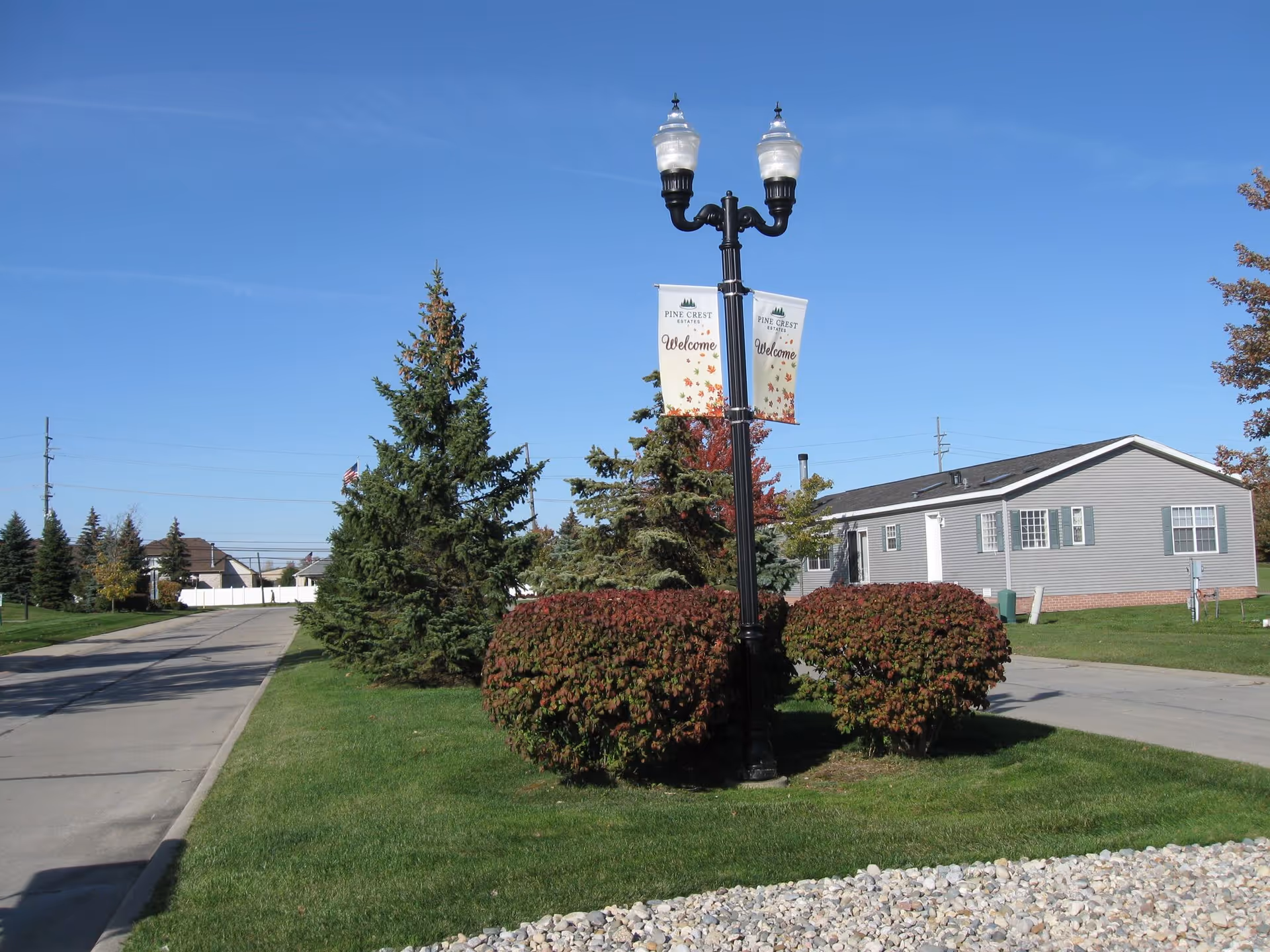 Outdoor view of Pine Crest Estates featuring a street lamp with two banners that say 'Pine Crest Estates Welcome', surrounded by trimmed bushes and pine trees, with a single-story gray building in the background under a clear blue sky.