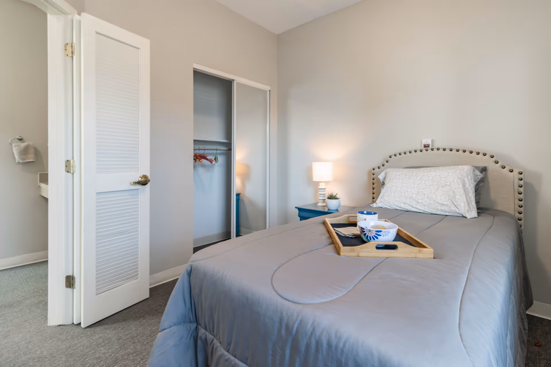 A bedroom with a neatly made bed covered in a gray comforter and a white pillow with a lace pattern. A wooden tray with a bowl and a cup is placed on the bed. To the right of the bed is a blue nightstand with a small lamp and a potted plant. Behind the bed is a light-colored wall with a small device mounted above the headboard. To the left, there is an open closet with sliding mirrored doors and an open door leading to a bathroom with a towel hanging on a ring.