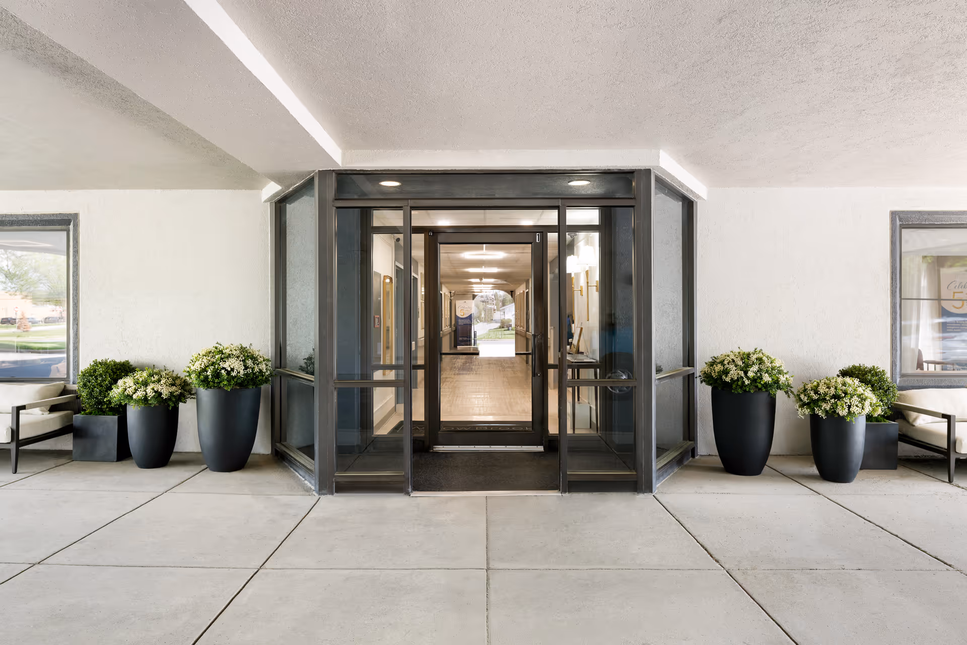 Covered building entrance with glass double doors, flanked by large black planters with greenery and seating.