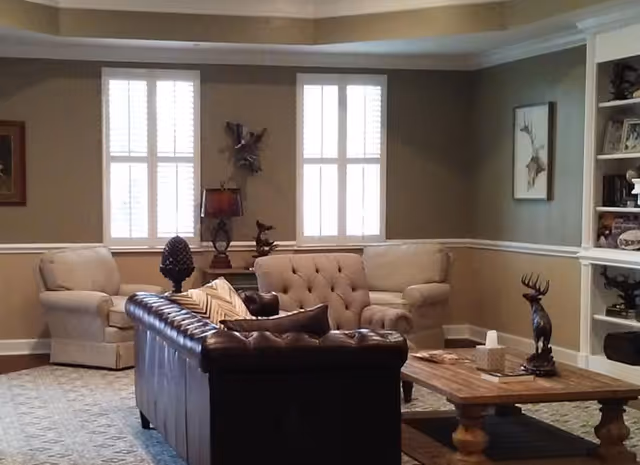 A cozy living room with a dark brown leather sofa, two beige armchairs, a wooden coffee table with decorative items including a tissue box and a deer figurine, beige walls with white trim, two windows with white shutters, a table lamp, and built-in shelves with various decorations.