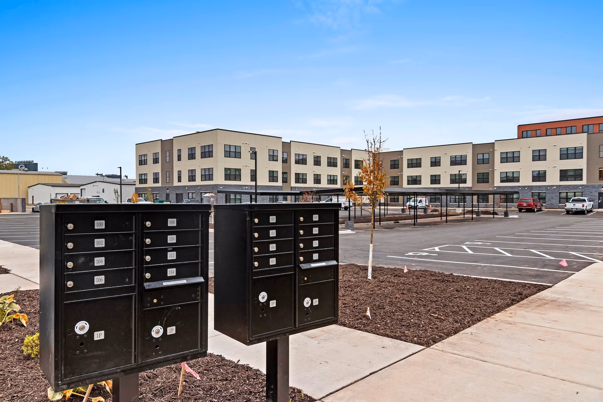 Two black community mailbox units on a sidewalk in front of a large three-story apartment building with a parking lot and young trees under a clear blue sky.