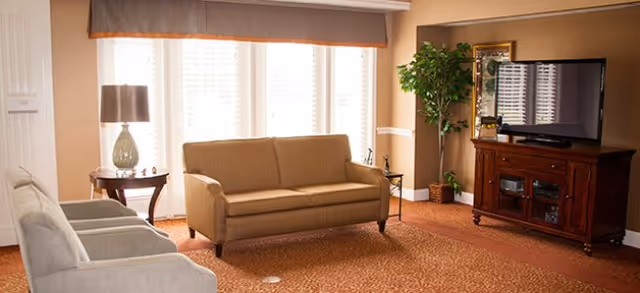A cozy living room with a beige sofa and two gray armchairs arranged around a wooden TV stand with a flat-screen television. A side table with a lamp is next to the sofa, and a large window with blinds and a valance lets in natural light. There is a green potted plant in the corner and a framed picture on the wall.