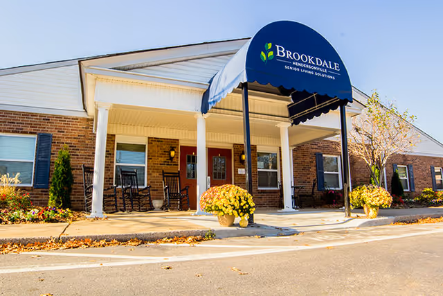 Front entrance of a senior living facility with a blue awning reading "Brookdale", a covered porch with rocking chairs and potted flowers.