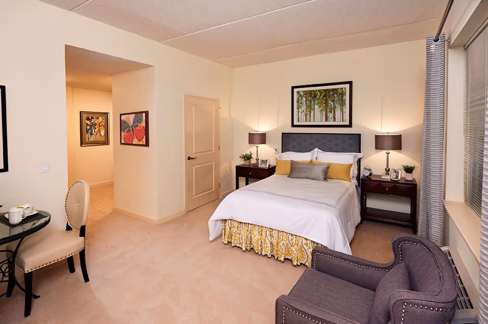 A cozy bedroom in an assisted living facility featuring a neatly made bed with white and yellow bedding, two bedside tables with lamps, a gray upholstered armchair, a small round table with a chair, and framed artwork on the walls. The room has beige carpeting and light-colored walls with a window covered by gray curtains.