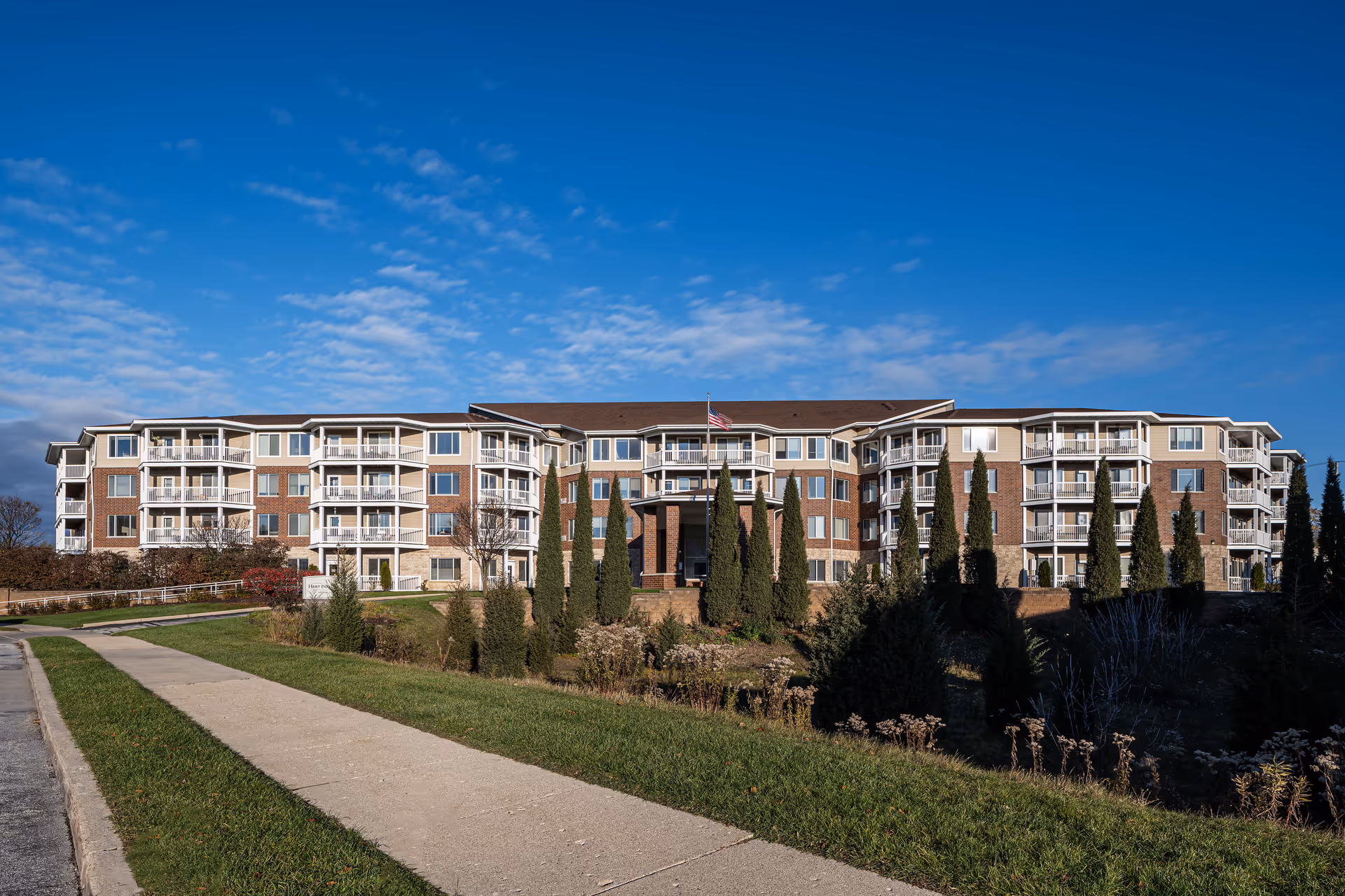 Exterior view of a large senior living facility building with multiple balconies, surrounded by landscaped greenery and tall trees under a blue sky with scattered clouds.