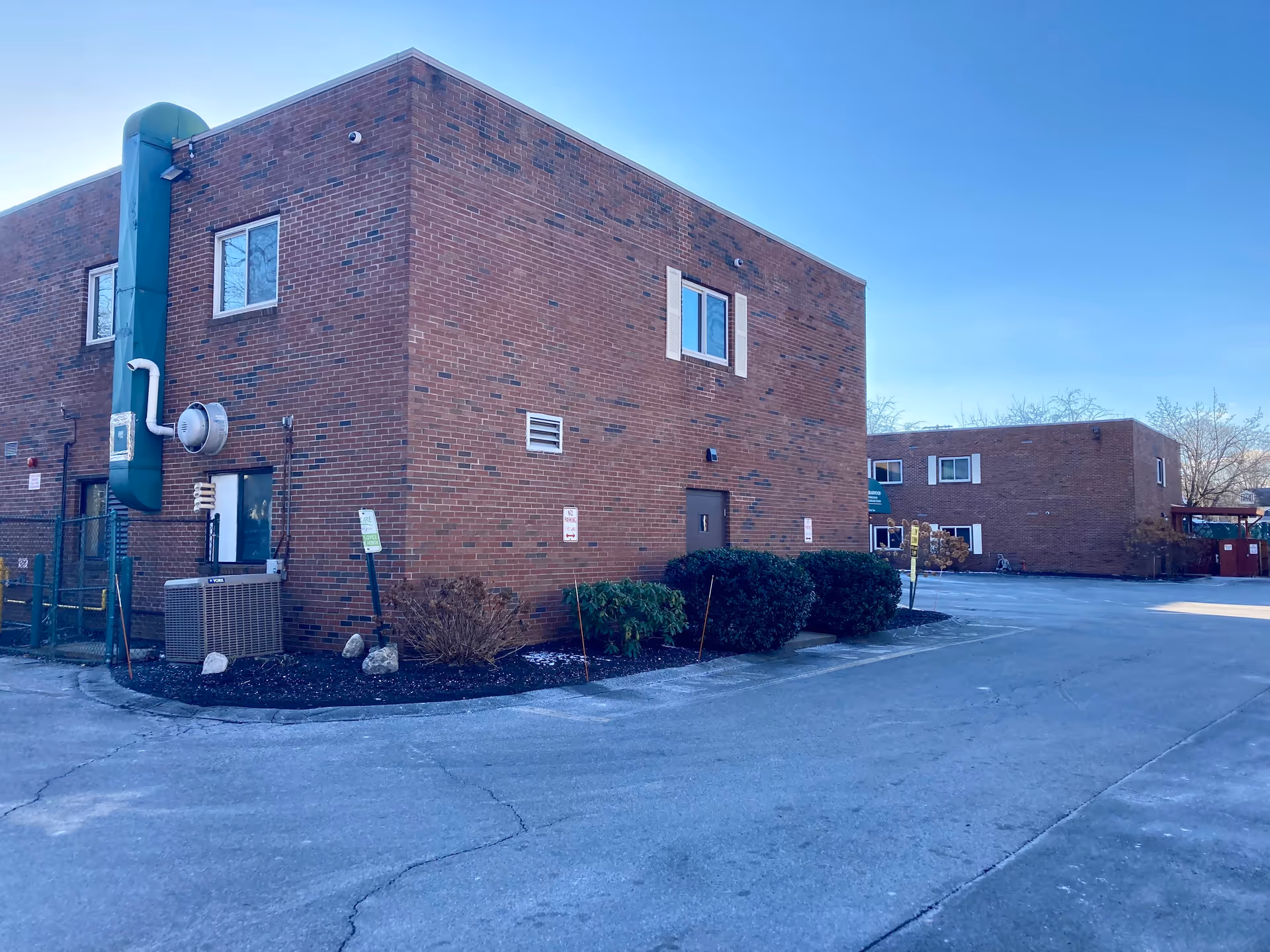Exterior view of a brick building with a flat roof, featuring windows and a door. The building is part of a facility with a paved parking area and some bushes along the side. The sky is clear and blue.
