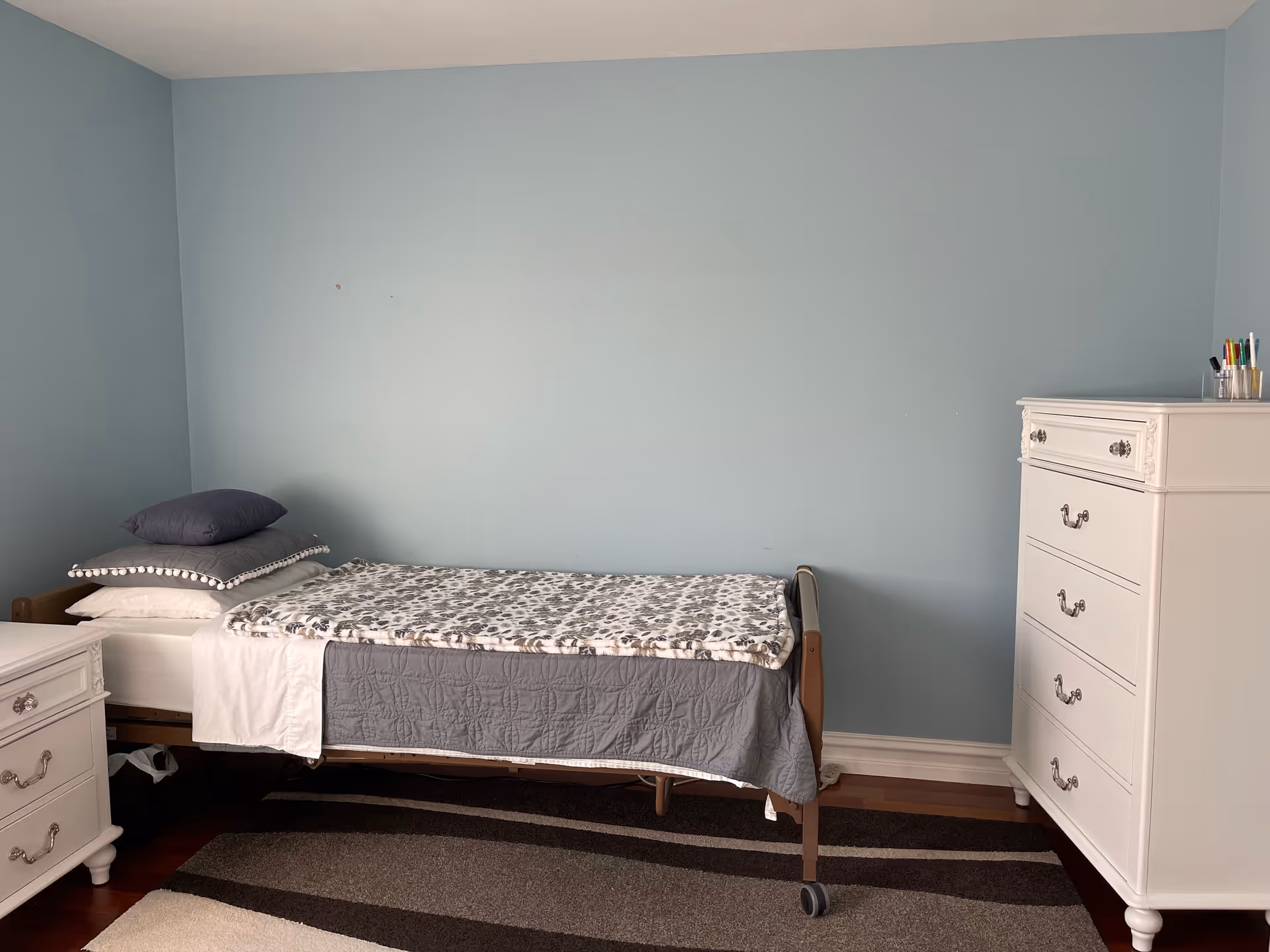A simple bedroom with a single bed against a light blue wall. The bed has a gray quilt and a patterned blanket on top, with several pillows stacked at the head. To the right of the bed is a white chest of drawers with decorative handles, and to the left is a matching white nightstand. The floor has a striped area rug in shades of brown and beige.