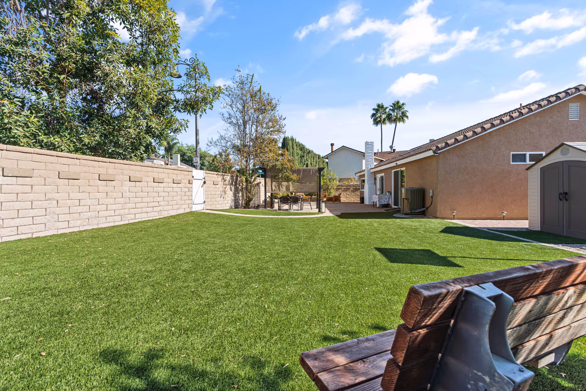 Outdoor area of Pacifica Home Care featuring a well-maintained green lawn, a wooden bench in the foreground, a patio area with a table and chairs under a canopy, surrounded by trees and a brick wall, with a clear blue sky above.