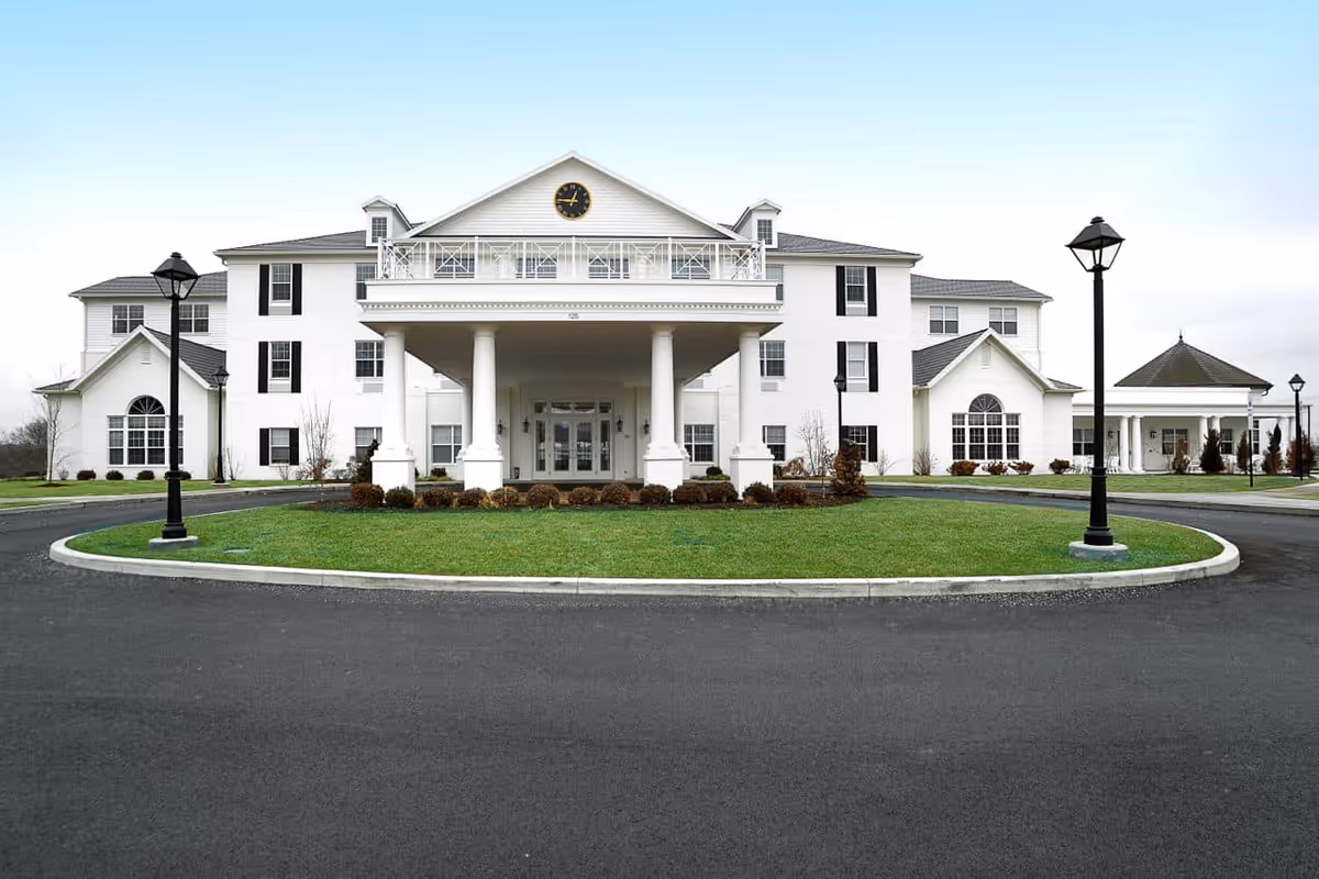 Front exterior view of a large, white senior living facility building with a covered entrance supported by columns, a clock above the entrance, and a circular driveway with green grass and lamp posts.