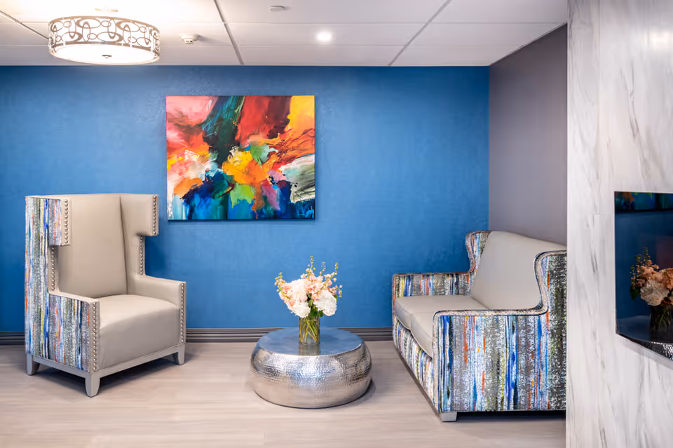 A modern sitting area with a beige armchair and a matching loveseat featuring colorful patterned sides. A round metallic coffee table with a vase of flowers is placed in the center. The background wall is painted blue and decorated with a vibrant abstract painting. A decorative ceiling light fixture is visible above.
