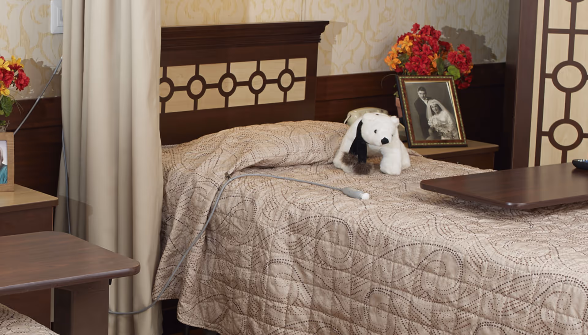 A neatly made single bed with a beige patterned quilt in a nursing home room. On the bed is a white stuffed dog toy and a remote control. Next to the bed is a wooden nightstand with a framed black and white wedding photo and a vase of red and orange flowers. The room has beige wallpaper and a curtain partially drawn to the side.