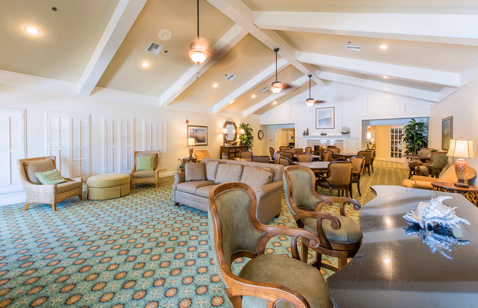 Bright, spacious senior living common room with sofas, armchairs, dining tables, patterned carpet and vaulted ceiling with fans.