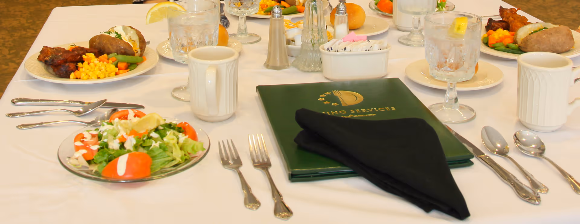 A dining table set with plates of food including salad, baked potato, corn, mixed vegetables, and meat. There are glasses of water with lemon, coffee mugs, silverware, salt and pepper shakers, and a green menu folder with a black napkin on top.