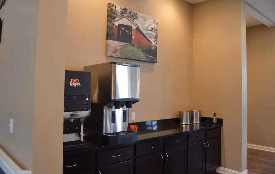 A beverage station in a senior living facility with a coffee machine, a water dispenser, and two metal containers on a black countertop with dark cabinets below. A framed picture of a red covered bridge hangs on the beige wall above the station.