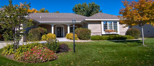 Single-story residential building with beige siding and a gray shingled roof, surrounded by green grass, bushes, and trees under a clear blue sky. A black lamppost stands in front of the building near a walkway leading to the entrance.