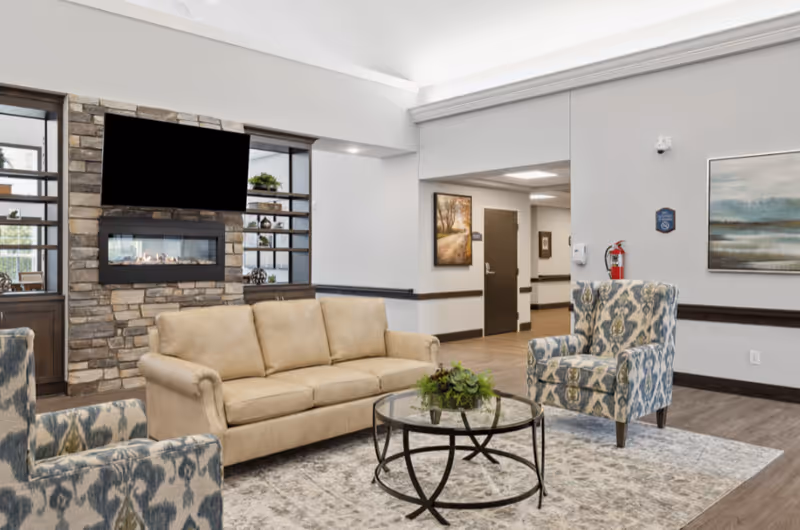 Lounge area with a beige sofa, patterned armchairs, round glass coffee table, and a wall-mounted TV above a stone fireplace.