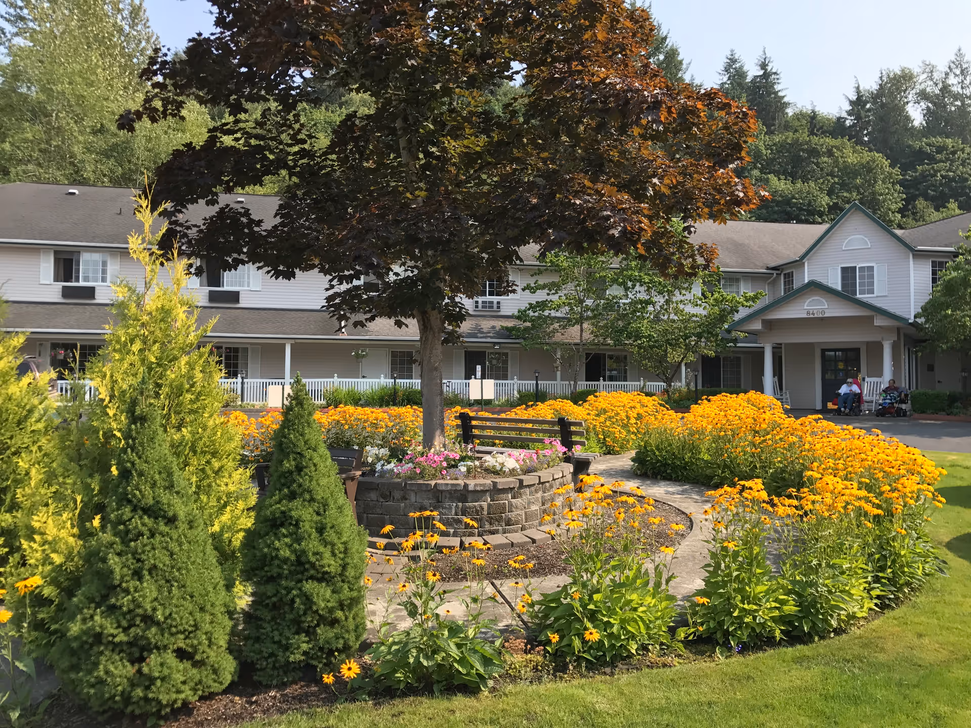 A senior living facility building with a well-maintained garden in front. The garden features a circular stone planter with a large tree in the center, surrounded by vibrant yellow flowers and green shrubs. There is a curved pathway and a bench near the planter. The building has two stories with white siding, multiple windows, and a covered entrance. Two people in wheelchairs are visible near the entrance.