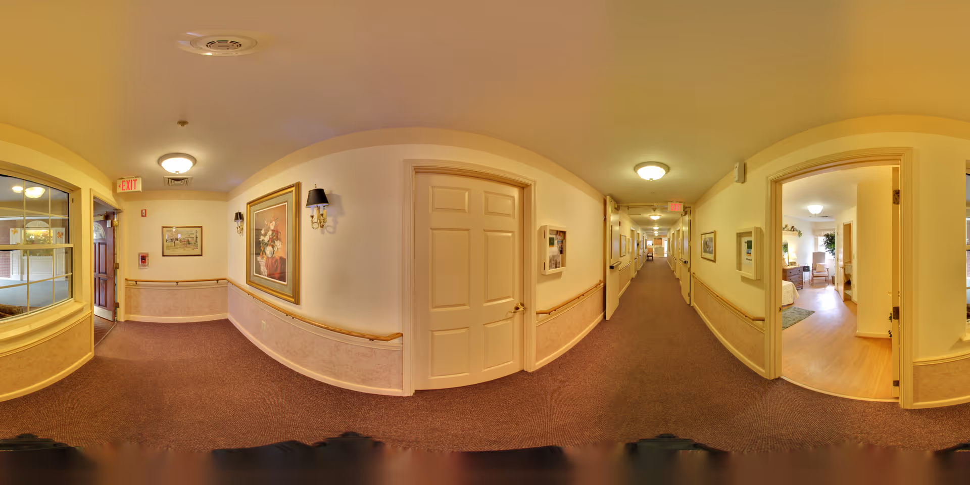 A long, well-lit hallway in a senior living facility with beige walls, carpeted floors, and several doors leading to rooms. The hallway is decorated with framed artwork and wall sconces. One room on the right side is open, showing a bedroom with wooden flooring, a bed, and a window with natural light.