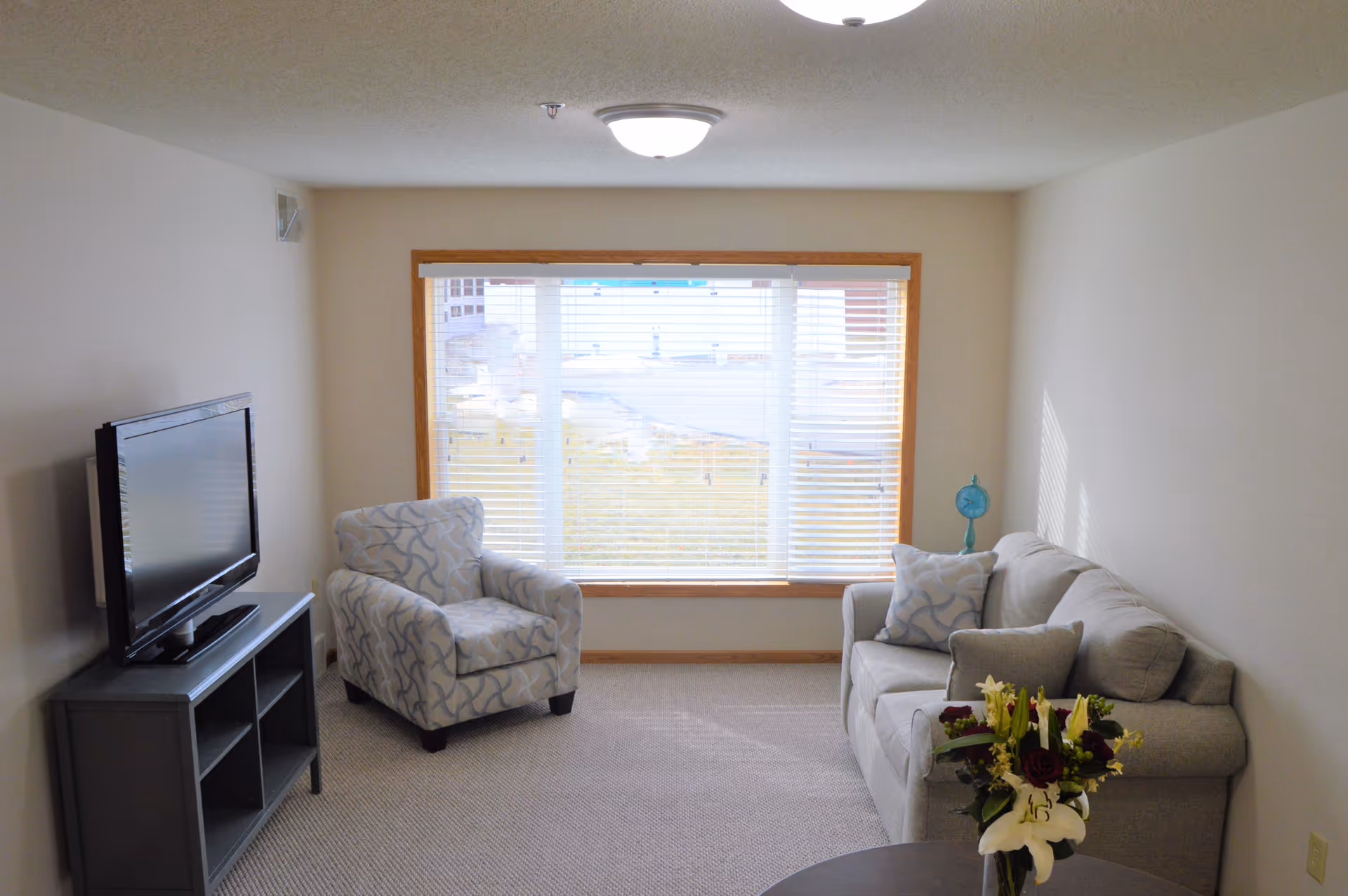 A small living room with a large window covered by white blinds. The room has a patterned armchair, a gray sofa with matching pillows, a small blue clock on a side table, a TV on a dark gray stand, and a round table with a bouquet of flowers in the foreground. The walls are light-colored and the floor is carpeted.