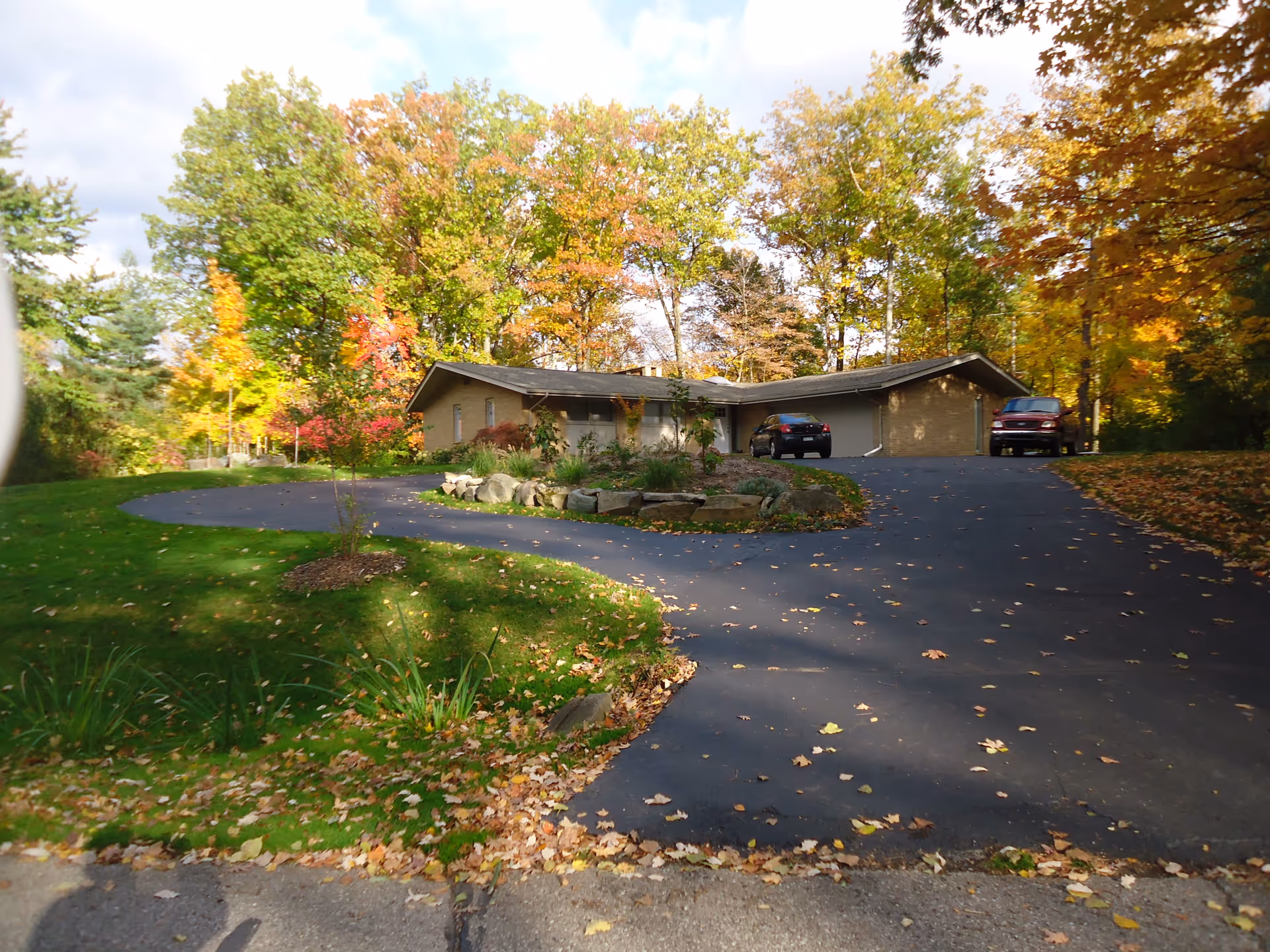A single-story building with a sloped roof surrounded by trees with autumn foliage. There is a curved asphalt driveway leading up to the building with two parked cars. The lawn is green with scattered fallen leaves.