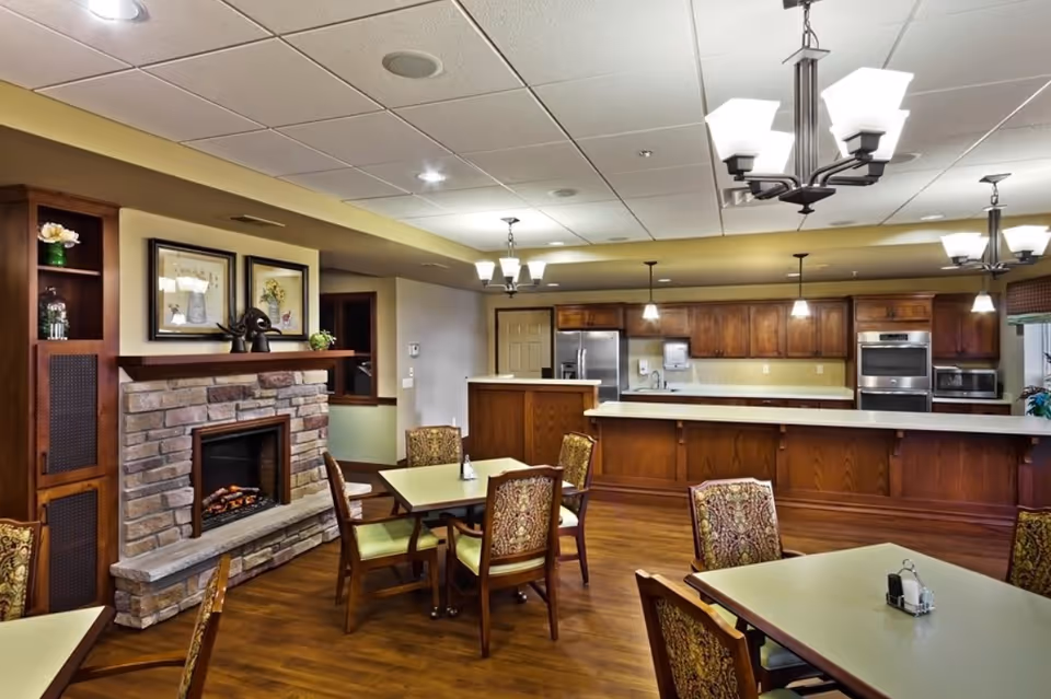 A spacious dining area with several tables and chairs featuring patterned upholstery. The room has a warm wooden floor and a stone fireplace with a wooden mantle decorated with framed artwork and a sculpture. In the background, there is a large kitchen area with wooden cabinets, a refrigerator, oven, microwave, and pendant lighting.
