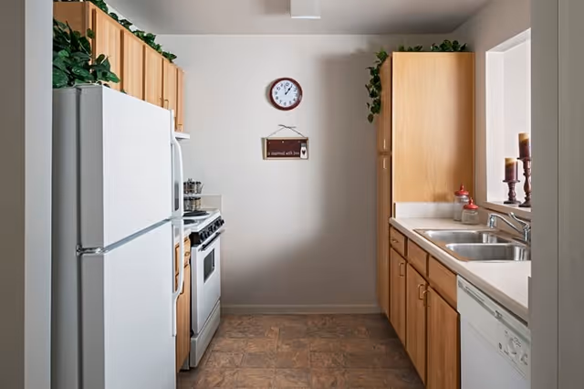 A small kitchen with light wood cabinets, a white refrigerator, a white stove, a double sink, and a dishwasher. There are decorative plants on top of the cabinets, a clock on the wall, and candles on the windowsill.