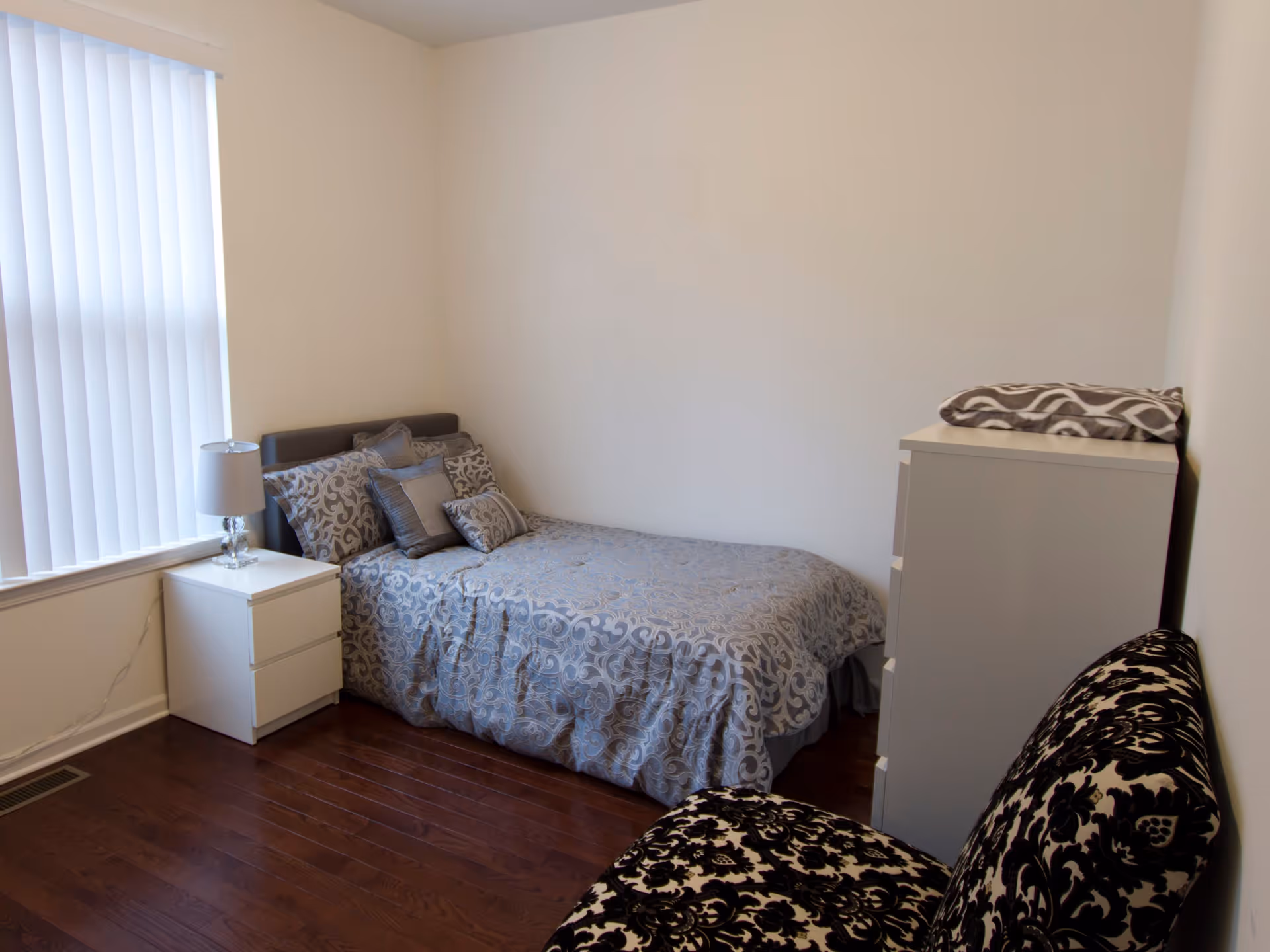 A small bedroom with a single bed covered in a gray patterned comforter and multiple pillows. Next to the bed is a white nightstand with a lamp. There is a white dresser with a folded blanket on top, and a black and white patterned chair in the foreground. The room has hardwood floors and a window with vertical blinds.