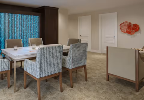 A modern dining area with a rectangular wooden table surrounded by six upholstered chairs with patterned fabric. The room has beige walls, carpeted floor, two white doors, and a decorative red wall art piece. A large dark wooden cabinet is visible against one wall.