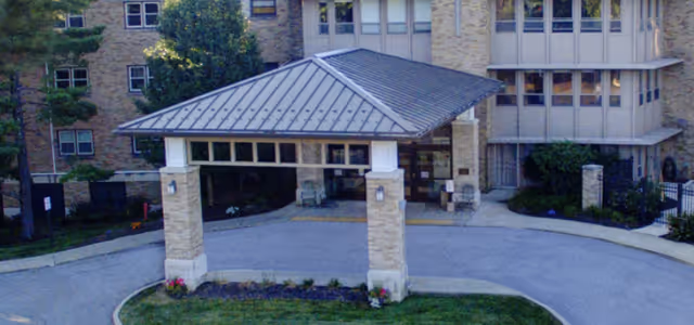 Front exterior view of The Village At St Edward at Fairlawn building entrance with a covered drop-off area supported by stone pillars, surrounded by greenery and a paved driveway.
