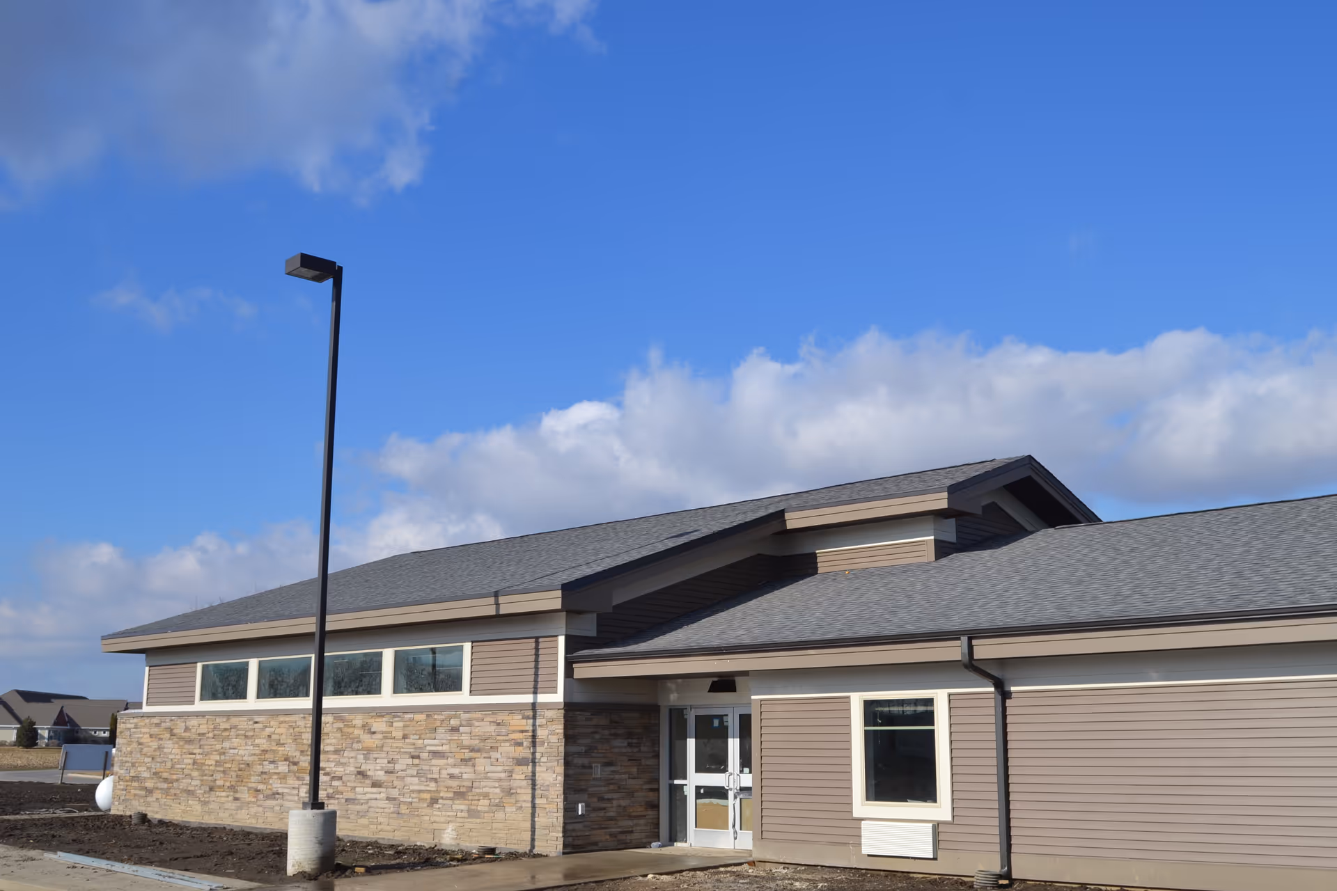 Exterior front of a single-story modern beige building with stone veneer, entrance doors, and a tall lamp post under a blue sky.