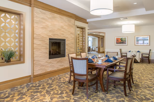 A dining room in a senior living facility with a wooden table set for six people with blue napkins and white cups. The room features patterned carpet, a modern fireplace with a textured wall, decorative wooden panels, and framed artwork on the walls. There are additional tables and chairs visible in the background under round ceiling lights.
