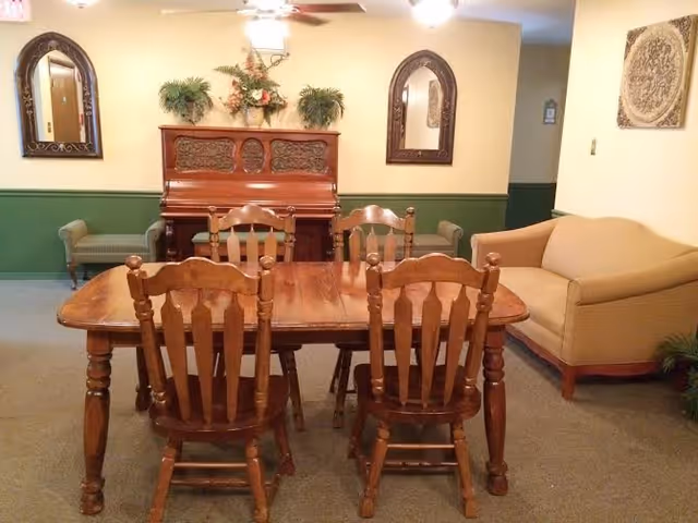 A cozy common room with a wooden table and chairs, an upright piano against the wall, and seating including a sofa and benches.