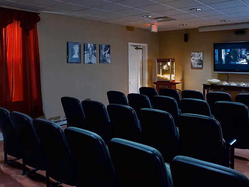 A small movie theater room with several rows of black cushioned seats facing a wall-mounted TV screen. There is a popcorn machine and bowls of popcorn on a table near the TV. The room has beige walls with black and white framed pictures and a red curtain covering a window.