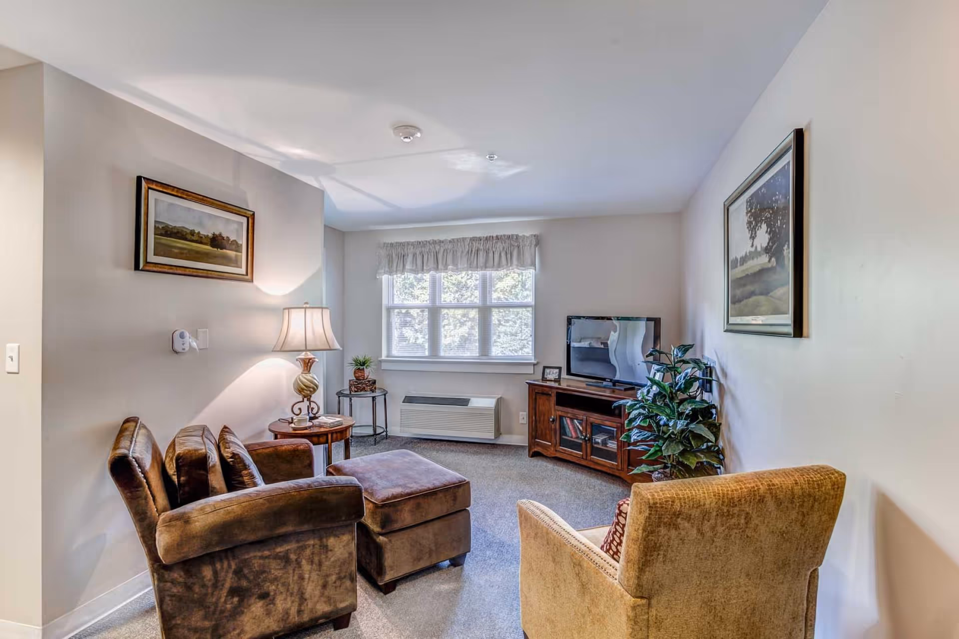 A cozy living room with a brown armchair and matching ottoman, a beige armchair, a wooden side table with a lamp, a small round table with a plant, a window with white curtains, a wooden TV stand with a flat-screen TV, a potted plant, and two framed landscape paintings on the walls.