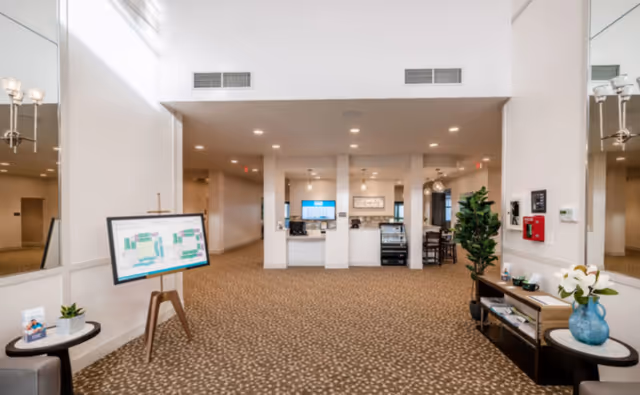 Spacious interior area of a senior living facility with patterned carpet, white walls, and recessed lighting. There is a large mirror on the left wall, a digital floor plan display on an easel, and a console table with decorative items and a plant on the right. In the background, there is a counter area with a computer and a display case, and seating is visible further back.