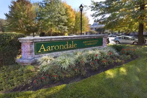 A brick sign reading Aarondale, surrounded by shrubs near a parking lot.