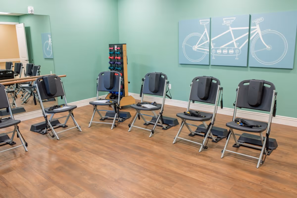 A small exercise room with six black folding chairs equipped with resistance bands and foot pedals arranged in a semicircle on a wooden floor. A rack of colorful dumbbells is positioned against a mint green wall, which is decorated with a three-panel artwork of a tandem bicycle. A large mirror covers part of one wall, reflecting the chairs and exercise equipment.